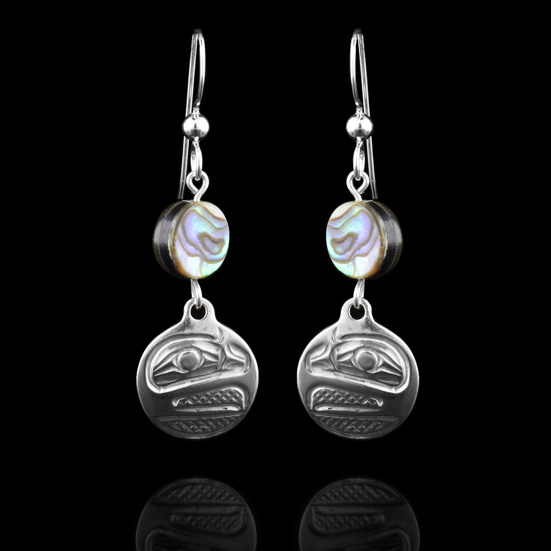 These dangling pearl earrings silver have small, circle hangs made from sterling silver. Below the hooks is a single, round abalone shell charm with the hangs dangling from them. On the hangs, the head of the Raven is shown at a side profile with its wide beak and eye detailed. There are cross hatching designs in the background of the portrait.