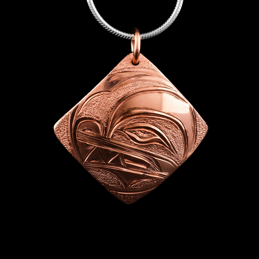 This Wolf pendant has a square, diamond shaped and is made from copper. The head of the Wolf is carved in a side profile in the center of the piece. The eye, nose and teeth are detailed and there are dimpled designs in the negative spaces of the scene.