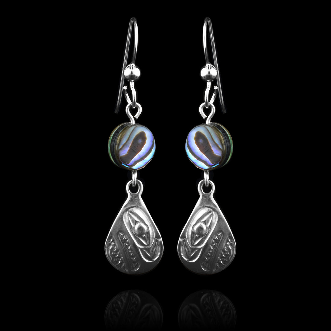 These abalone drop earrings silver have long teardrop shaped hangs made from sterling silver. Below the hooks is a single, round abalone shell charm with the hangs dangling from them. On the hangs, the head of the Raven is shown at a side profile with its wide beak and eyes detailed. There are cross hatching designs in the background of the portrait.