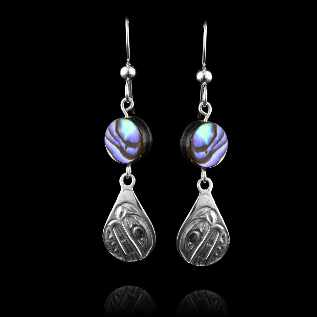 These abalone drop earrings silver have small teardrop shaped hangs made from sterling silver. Below the hooks is a single, round abalone shell charm with the hangs dangling from them. On the hangs, the head of the Orca is shown at a side profile with its teeth and eyes detailed. There are cross hatching designs in the background of the portrait.