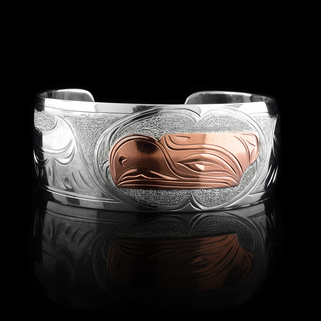 This Eagle bracelet has a single, wide band with a gap in the back and the head of the Eagle is depicted in the center of it. The head is done in copper and at a side profile. On both side of the band, there are carvings showing the wings outstretched across the band with feather details.