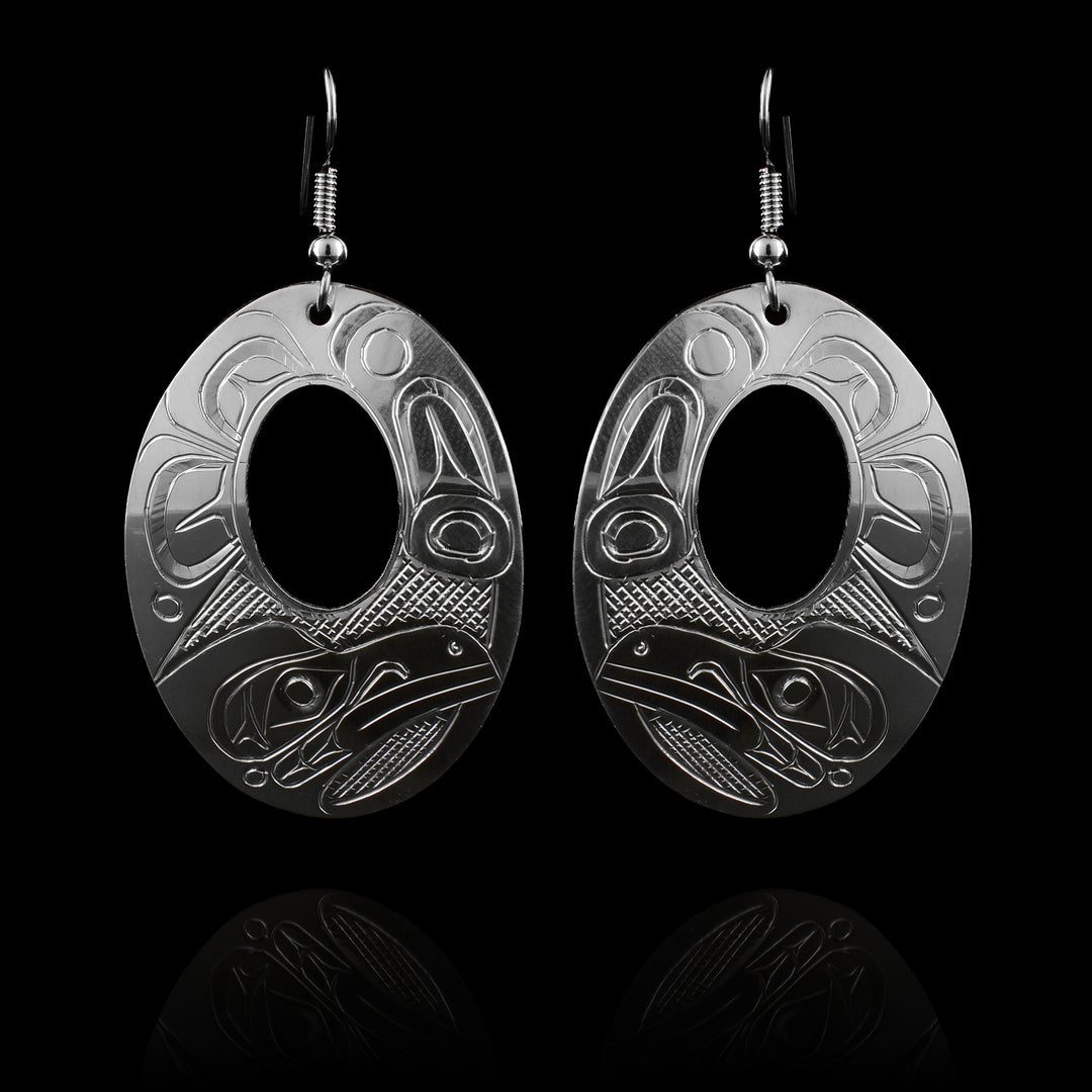 These large silver earrings have wide, oval shaped hangs with small, oval cutouts in the center of them. The head of the Raven is carved into the bottom with a side profile. There are feather designs made around the rest of the piece.