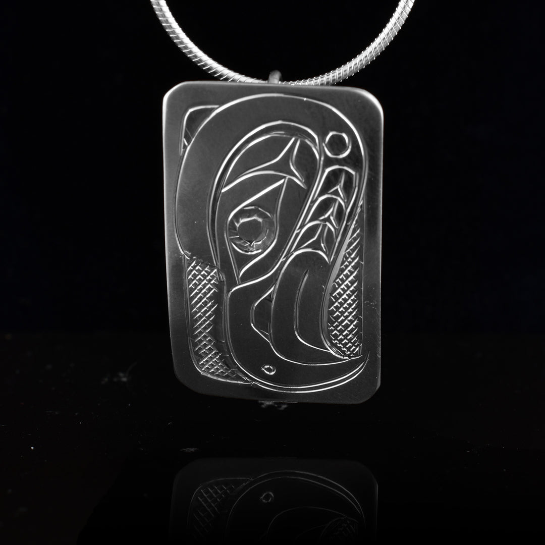 This Eagle head pendant is made from sterling silver and has a small rectangle shape. There is a depiction of the head of the Eagle carved in a side profile on the piece. Its beak is facing down and there are designs on its face. There are cross hatching designs in the background of the scene.