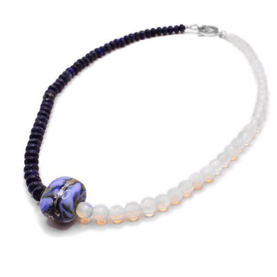 This opalite necklace depicts dark blue lapis beads on one side and opalite beads on the other. The centerpiece is a large, asymmetrical glass bead with the colours light blue, gold, and green. 