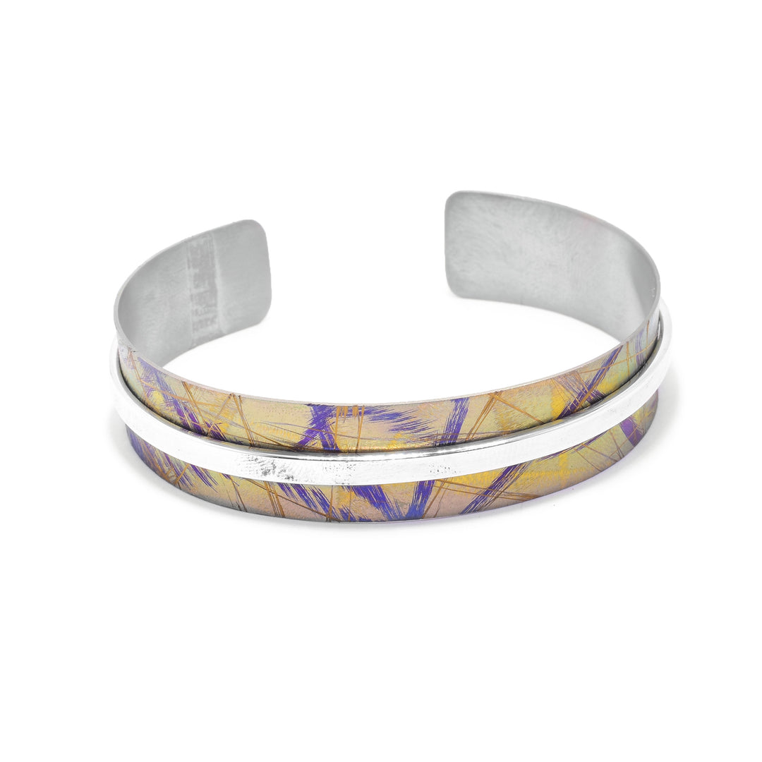 This titanium bangle bracelet has a titanium band with a gap in the back. There are yellow, blue and gold designs on the titanium. There is a thin, sterling silver band going around the piece.