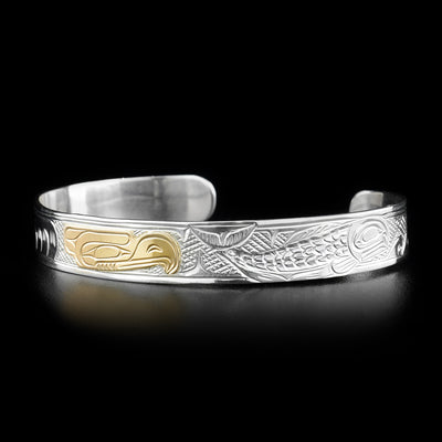 This eagle bracelet depicts a gold eagle head in the center facing a full-bodied salmon to its right. Waves have been carved in front of the salmon. Background has been cross-hatched.