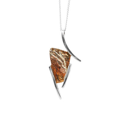 This oxidized silver necklace is in an abstract shape with 3 silver lines encasing a rectangular oxidized piece of silver that is orange and brown in colour.