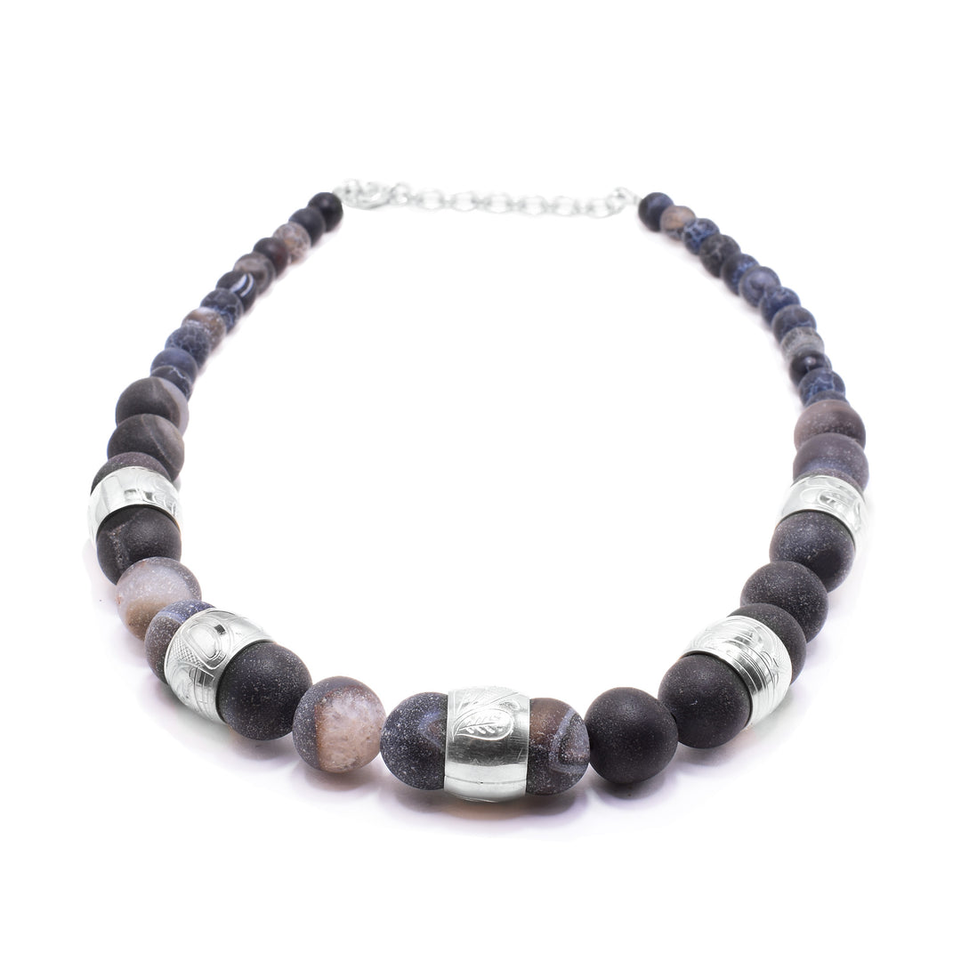 This chunky dark agate necklace has sphere shaped gemstone beads that are larger at the bottom portion of the piece and smaller at the tops of it. There are five sterling silver spirit beads that are ditributef through the large beads. The spirit beads have depictions of the Bear, the Orca, the Hummingbird, the Sun and the Frog carved into the surface of them.