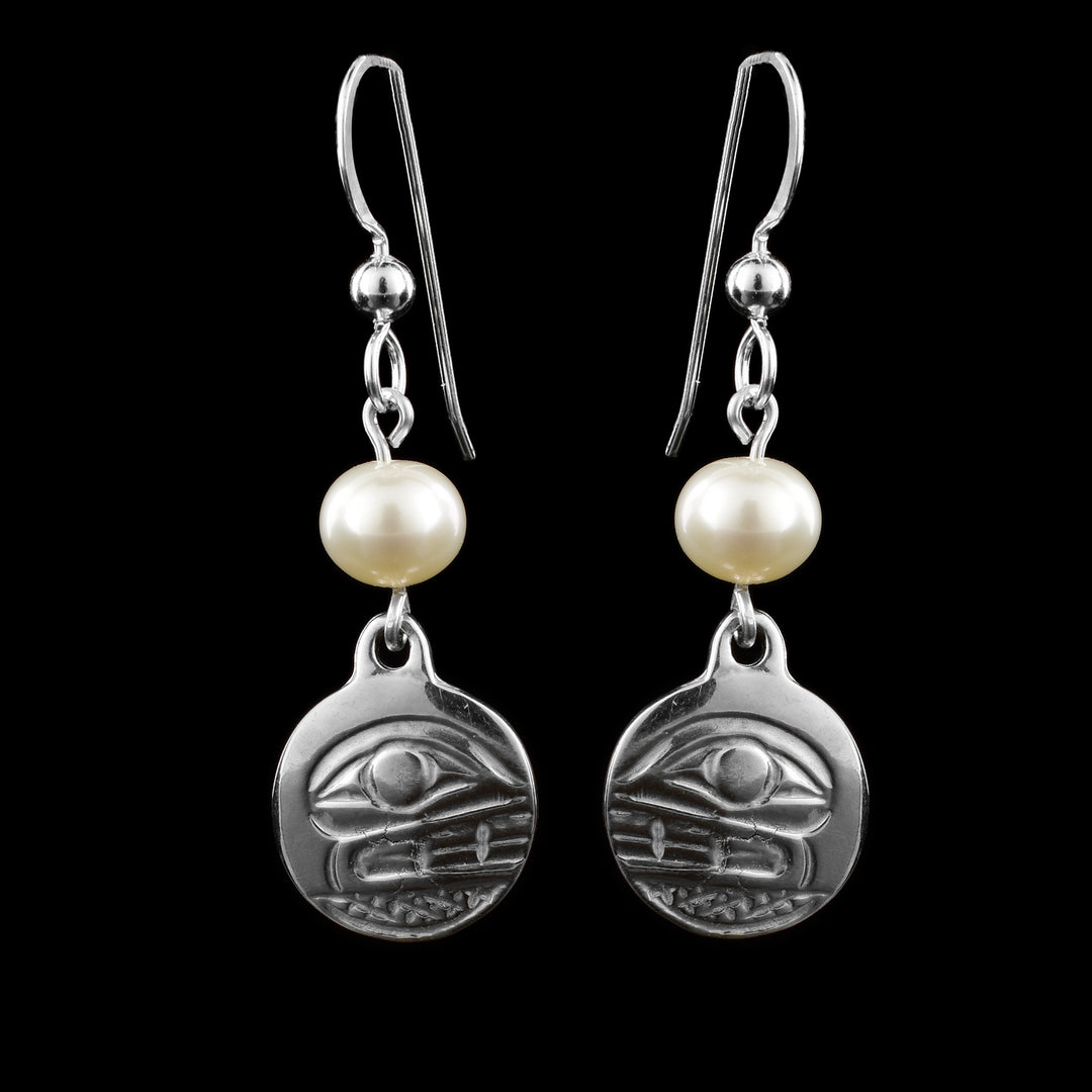 These dangling pearl earrings silver have small, circle hangs made from sterling silver. Below the hooks is a single, white pearl with the hangs dangling from them. On the hangs, the head of the Orca is shown at a side profile with its eye and teeth detailed. There are cross hatching designs in the background of the portrait.