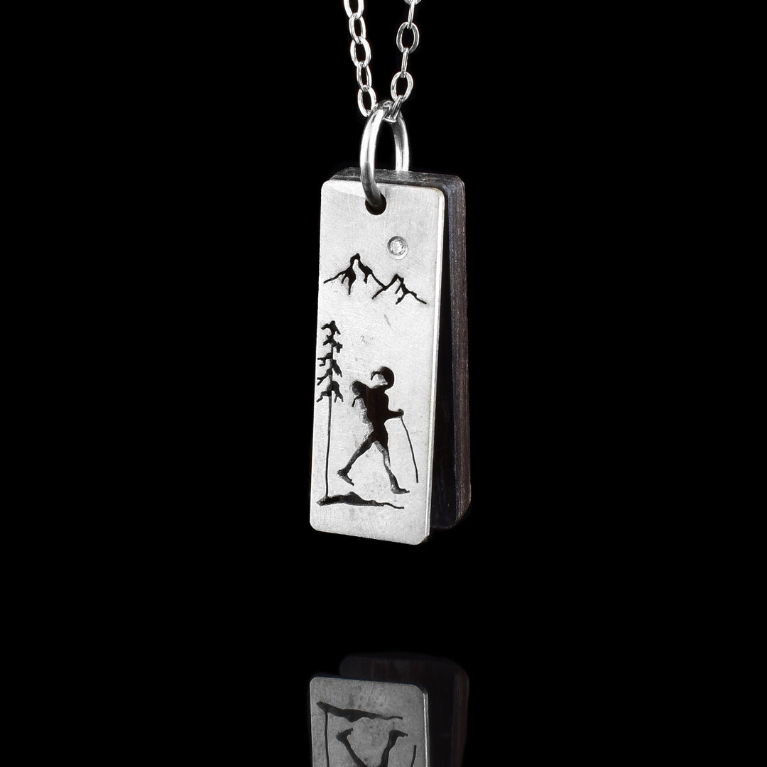 This hiker mountain pendant has a sterling silver rectangle pendant with a cutout pattern on it. There is a silhouette of a hiker in the bottom with a tree reaching up above him. At the top of the pendant there are two mountain peaks. There is a dark, brown wooden piece behind the pendant that matches it in size.
