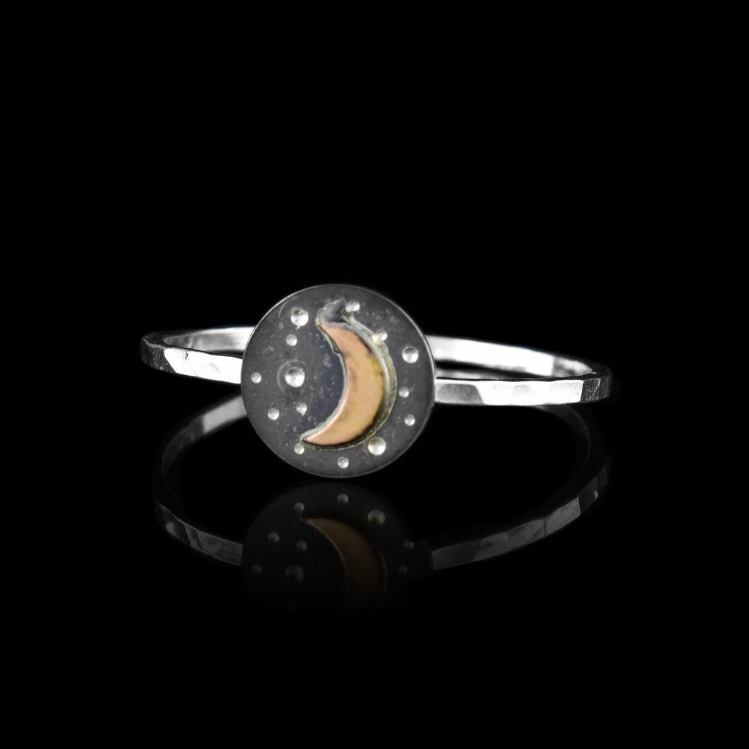 This crescent moon ring has a thin, sterling silver band with a silver circle on top of it. There are dimples made on the circle representing stars and there is a crescent moon made from 14K gold filled material in the center of the piece.