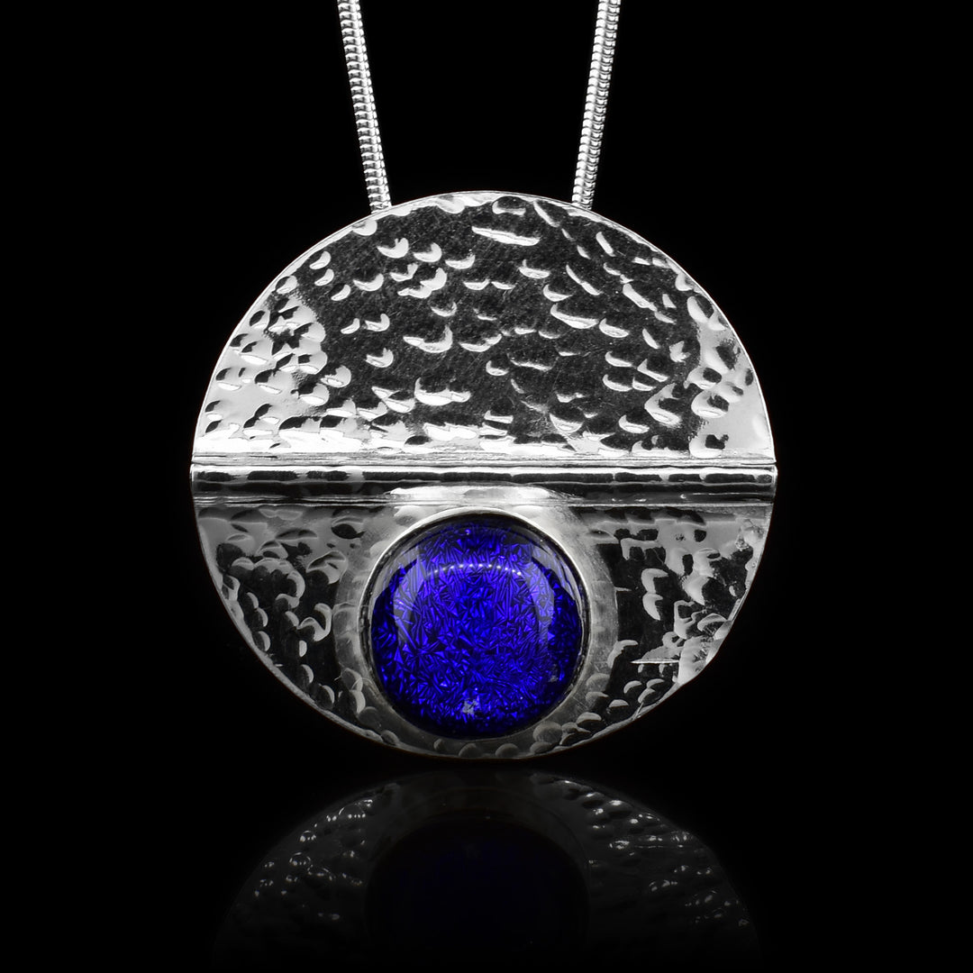 This sterling silver circle pendant has a dark blue colored fused glass bead embossed in the bottom half of the piece. There is a straight crease made horizontally through the piece and there are dimpled textures made all across the piece.