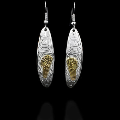 Each dangle earring consists of a long, curved, oval featuring a hummingbird pointing downwards with beak in flower. Face done in 14K yellow gold. Cross-hatching background. Hand-carved by Kwakwaka’wakw artist William Cook.