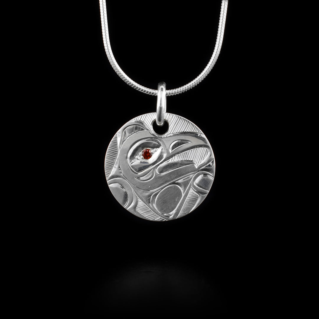 This Sterling Silver Eagle Pendant with Garnet is hand carved by artist, Hollie Bartlett. The piece is a sterling silver circle and the Eagle is depicted in side view on it. The head of the eagle is in the center and its wing is bent around it at the bottom of the piece. There is a red, garnet gem embedded in the eye of the legend.