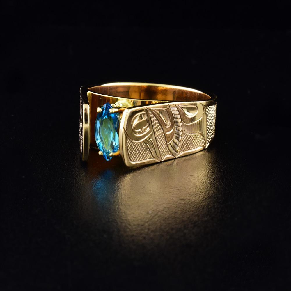 This blue topaz ring gold has a 14K gold band and London blue topaz gem mounted in the center. The Hummingbird is carved into the sides of the band. Its is shown in a side profile with its head looking upwards and its wings at its side. The band extends up, above the gem in a cathedral fashion.