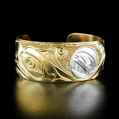 Brass and Silver 1" Orca and Thunderbird Bracelet by Paddy Seaweed. The design depicts a profile of an orca's face in the center of the bracelet made out of sterling silver. There are two profiles of thunderbirds faces facing away from the orca made from brass. The background has been delicately hand carved to allow for the legends to stand out.