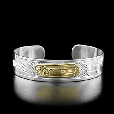 Sterling Silver and 14K Gold 1/2" Raven Bracelet by Victoria Harper. In the center of the bracelet the artist has hand-carved the profile of a raven's head looking towards the right out of 14k gold. The raven has a short beak and a feather on the back of its head. On both sides of the bracelet the artist has hand-carved intricate designs out of sterling silver to represent the raven's body and feathers.