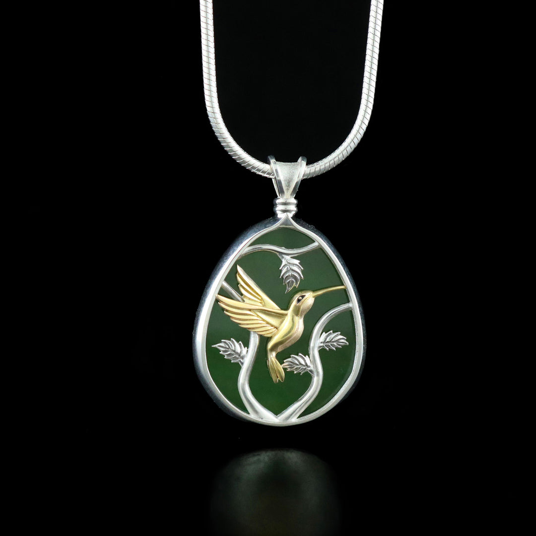 This jade pendant necklace has an oval shaped with a depiction of a Hummingbird flying around a tree. The hummingbird is made from 14K gold and the jade and three designs are set in sterling silver.