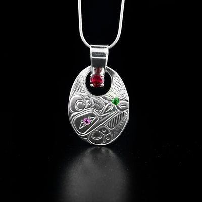 Dazzling oval cut out hummingbird pendant hand-carved by Coast Salish and Cree artist Richard Lang. Made of sterling silver and lab-created stones. Richard has set a pink stone in the eye of the hummingbird, a green stone in the center of the flower and a red stone at the bottom of the bail. Pendant measures 1.75" x 1" including bail. Chain not included.