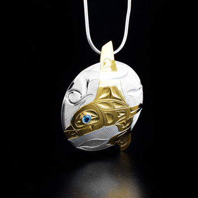 Stunning Silver and Gold Orca Pendant with Blue Topaz hand-carved by Tlingit artist Fred Myra. He carved the sterling silver piece first and then applied 18K gold plating on it. There is a blue topaz set in the orca's eye. Pendant measures 1.8" x 1.4". Hidden bail on back. Chain not included.