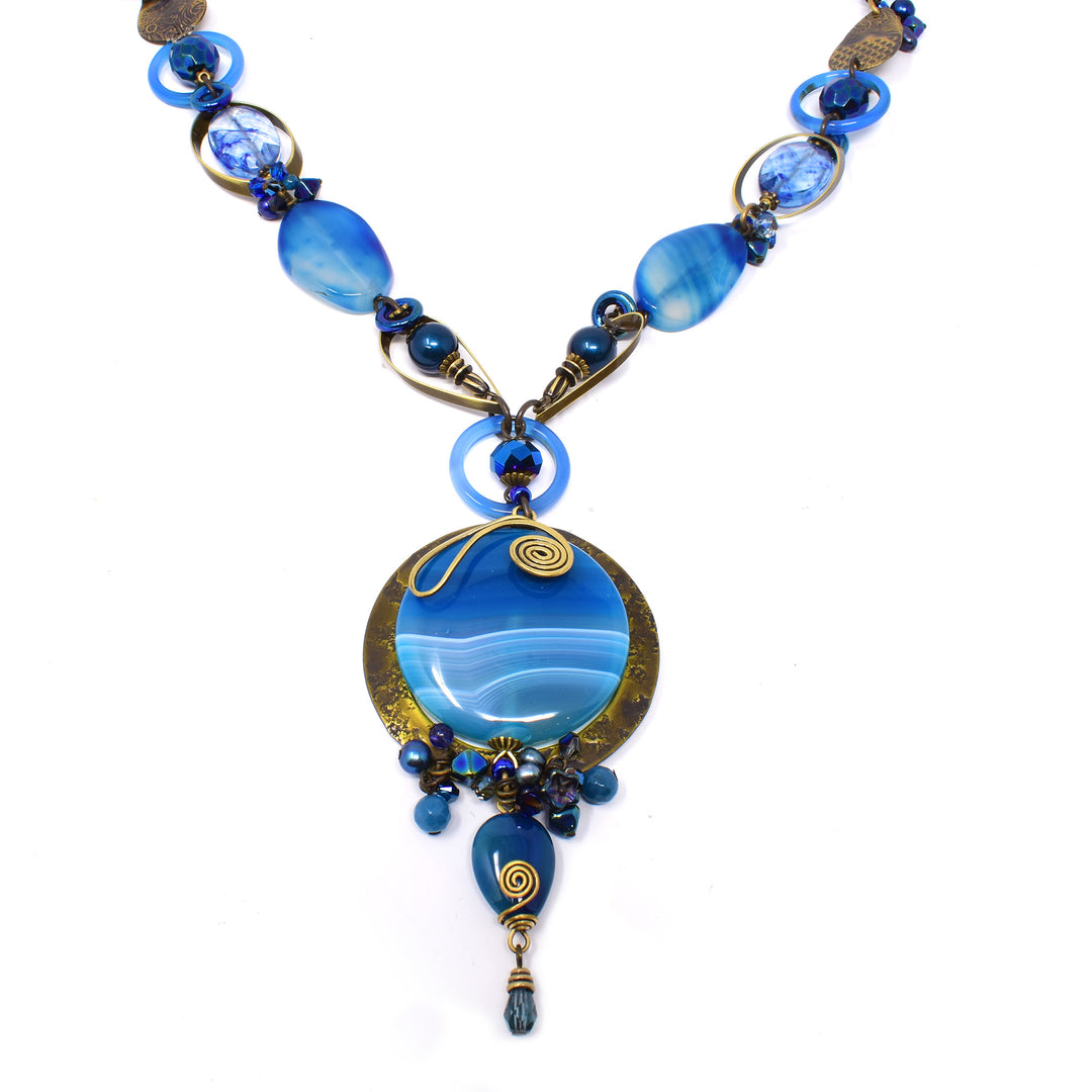 This gorgeous blue gemstone necklace is made from hand worked brass, lampworked glass, freshwater pearls, Austrian crystal, dyed agate, dyed quartz, glass and kyanite. There is a large, circle pendant with wave patterns on it and crystal and brass charms around it. There are crystal, gemstone and brass beads of different shapes, sizes and pattern running along the entire piece.
