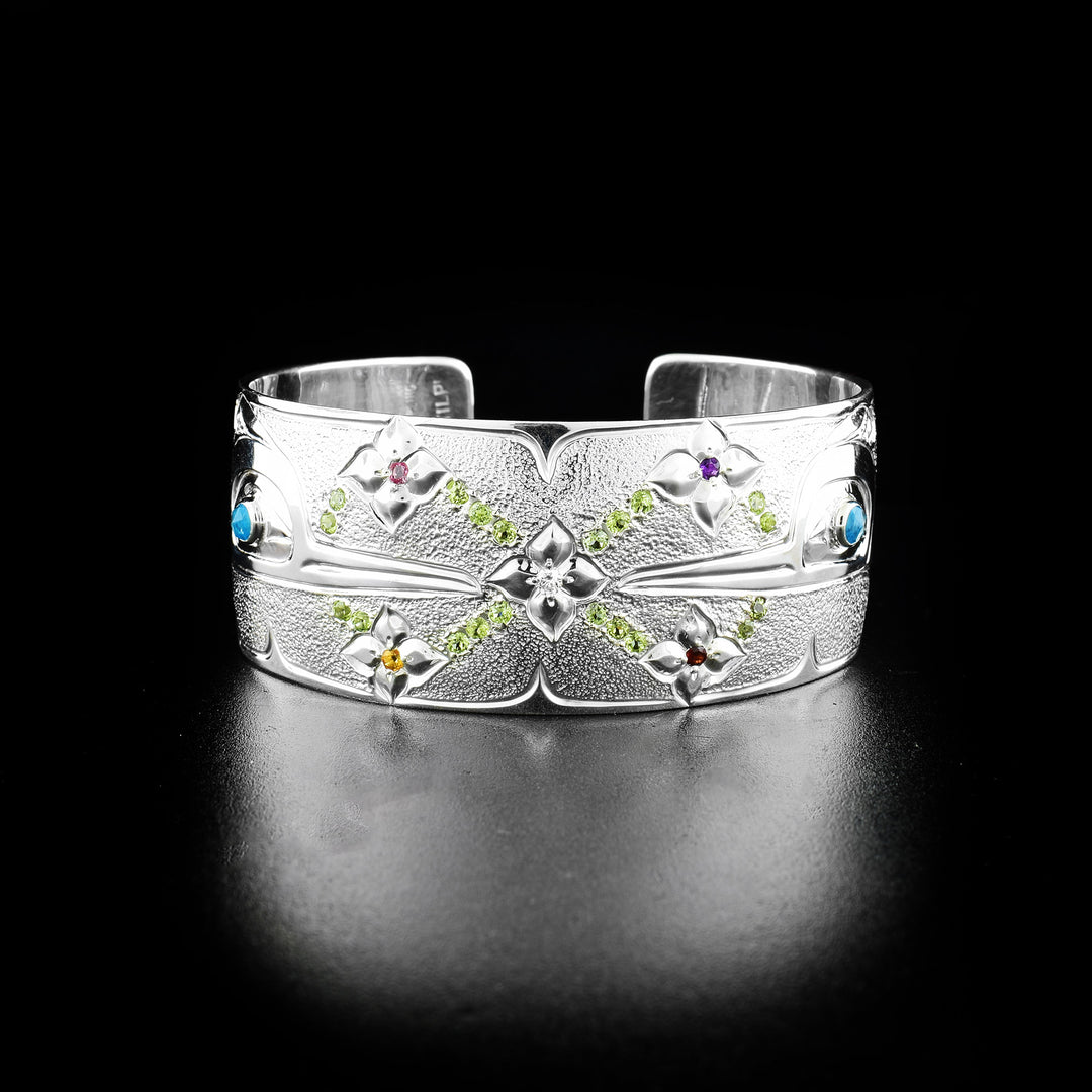 This Hummingbird bracelet cuff is made from sterling silver, peridot, amethyst, blue topaz, diamond, garnet and citrine. The piece has a single, wide band with a gap in the back. There are depictions of the Hummingbird carved into the band and the gems are imbedded into the band in designs.