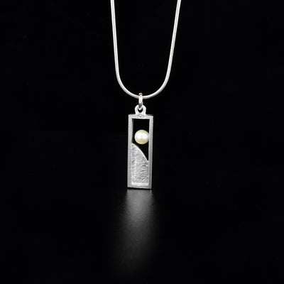 Sterling Silver Window Pearl Necklace by Jean Bastien. The pendant is in a rectangular shape. The bottom portion of the pendant has been filled in with sterling silver. The artist has included a white freshwater pearl towards the top of the necklace and has attached it to one of the sides of the pendant to give it a "floating effect". 