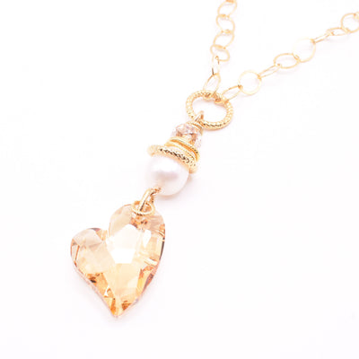 Swarovski Crystal Heart and Pearl Pendant Necklace by Karley Smith. At the end of the necklace is a Swarovski crystal heart in an orangey colour. Above the heart is a white freshwater pearl. The artist has added gold-plated accents that lay on top of the freshwater pearl. On top of the gold-plated accents is a small Swarovski crystal and finally, a large hoop that connects the gold-filled chain together.