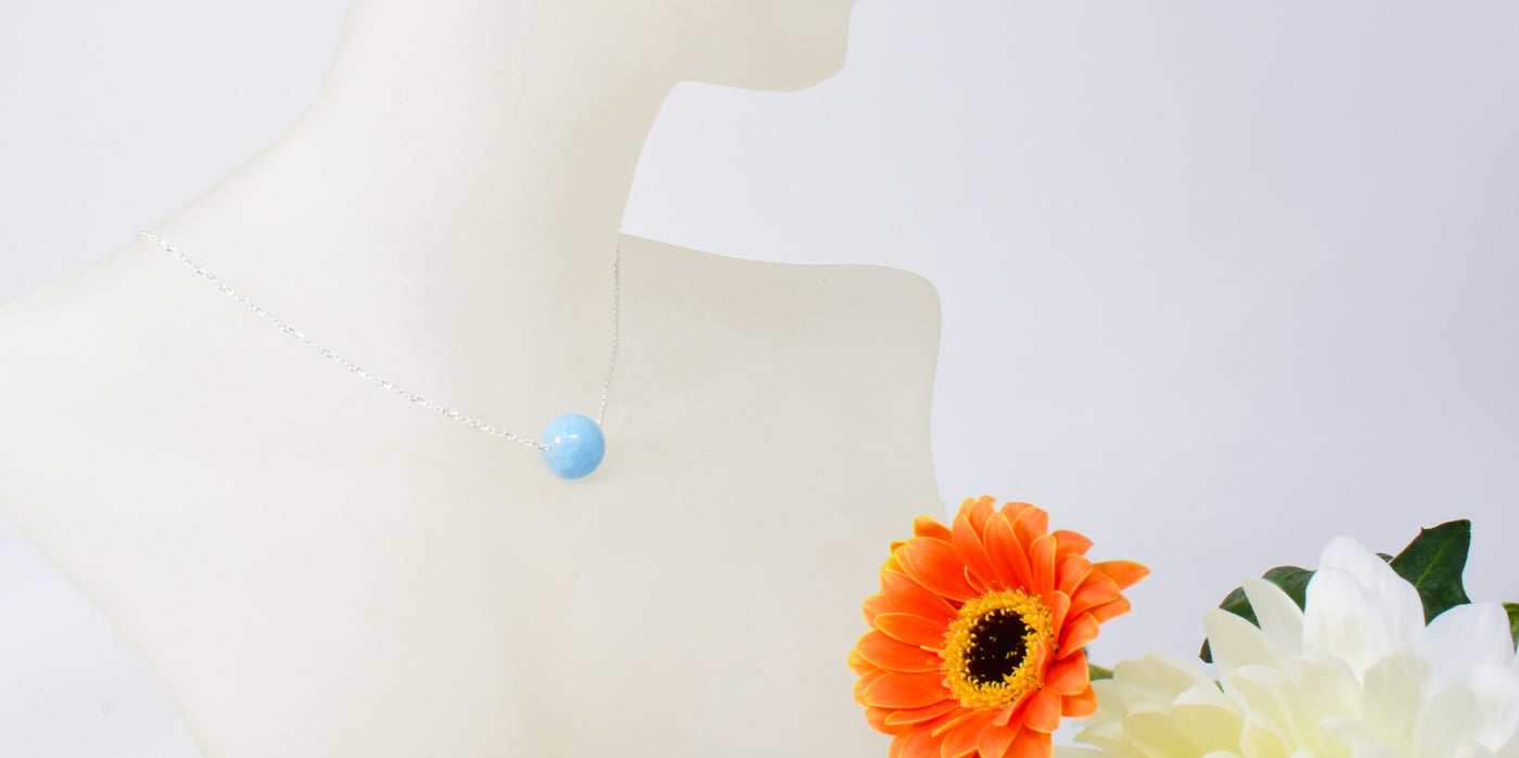 A mannequin models a floating Aquamarine necklace with a silver link chain. Summer flowers offset the image.