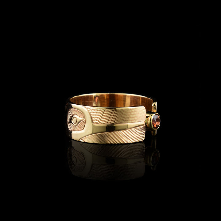 This 14K gold garnet ring has a wide band made from gold and the band is split in the center of the band. There is a small garnet gem held in the center of the split in the band. There are carvings on the surface of the band with a head of the Hummingbird at a side profile on both sides of the gem. The beak extends to the end of the band and there are intricate hatching designs in the background of the scene.