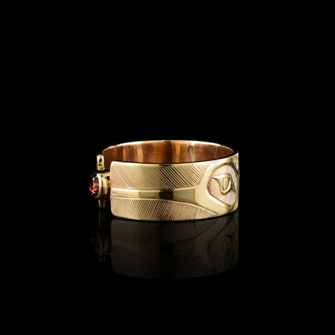 This 14K gold garnet ring has a wide band made from gold and the band is split in the center of the band. There is a small garnet gem held in the center of the split in the band. There are carvings on the surface of the band with a head of the Hummingbird at a side profile on both sides of the gem. The beak extends to the end of the band and there are intricate hatching designs in the background of the scene.