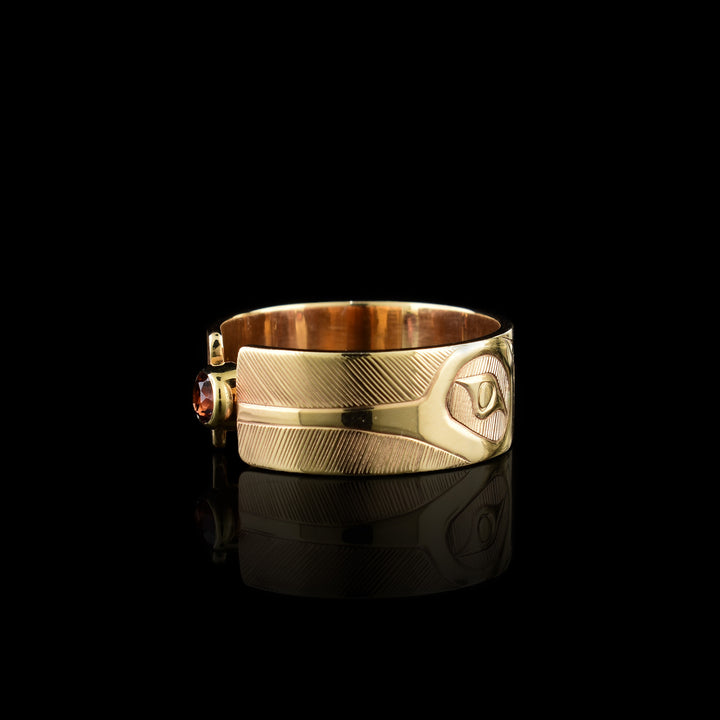 This 14K gold garnet ring has a wide band made from gold and the band is split in the center of the band. There is a small garnet gem held in the center of the split in the band. There are carvings on the surface of the band with a head of the Hummingbird at a side profile on both sides of the gem. The beak extends to the end of the band and there are intricate hatching designs in the background of the scene.