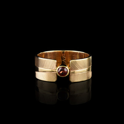 This 14K gold garnet ring has a wide band made from gold and the band is split in the center of the band. There is a small garnet gem held in the center of the split in the band. There are carvings on the surface of the band with a head of the Hummingbird at a side profile on both sides of the gem. The beak extends to the end of the band and there are intricate hatching designs in the background of the scene.
