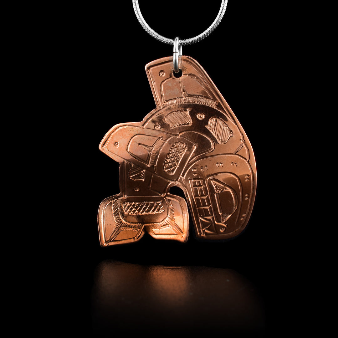 This Orca whale pendant is made from copper and is shaped like a breaching Orca. The Orca's body is curved with its head and tail at the bottom of the piece and its dorsal fin at the top. In has a fin on its side that extends out towards the top. There are carvings on the surface of the piece showing its eye and teeth. There are also designs that detail the rest of the Orca.