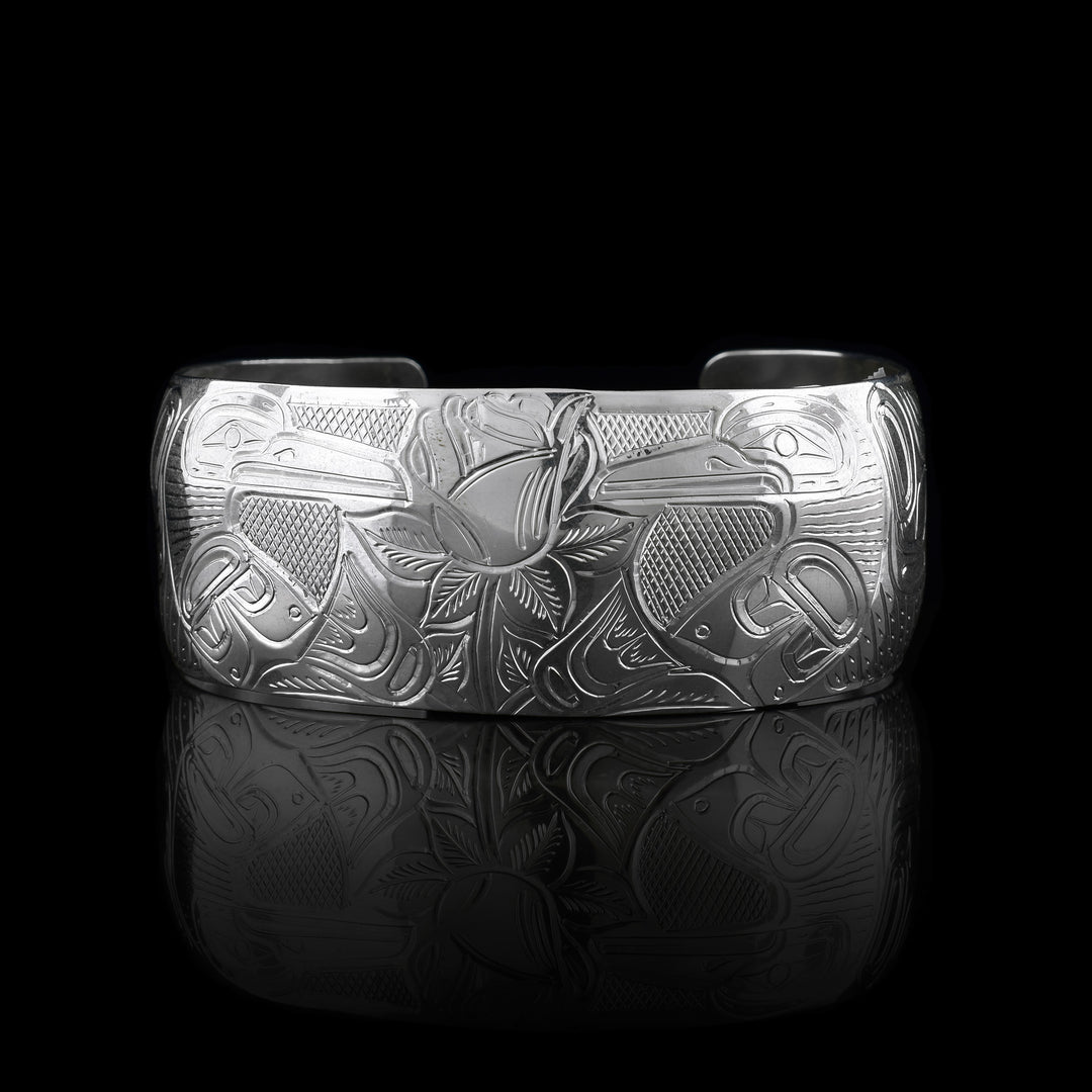 This Hummingbird and flower cuff bracelet is made from sterling silver and has a wide band with a gap in the back. There are two Hummingbirds at a side prolife facing a rose with leaves carved into the center of the piece. There are designs and cross hatching around the scene.