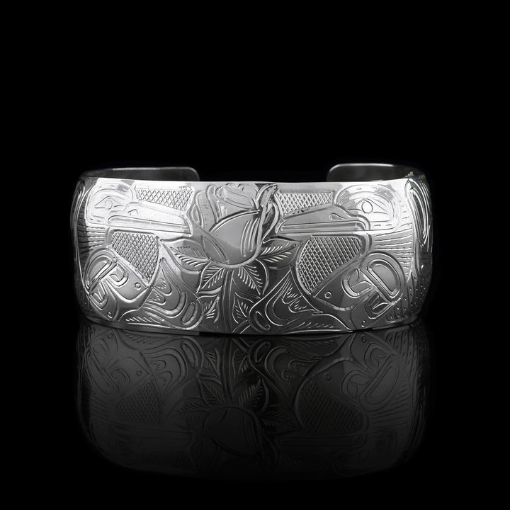 This Hummingbird and flower cuff bracelet is made from sterling silver and has a wide band with a gap in the back. There are two Hummingbirds at a side prolife facing a rose with leaves carved into the center of the piece. There are designs and cross hatching around the scene.