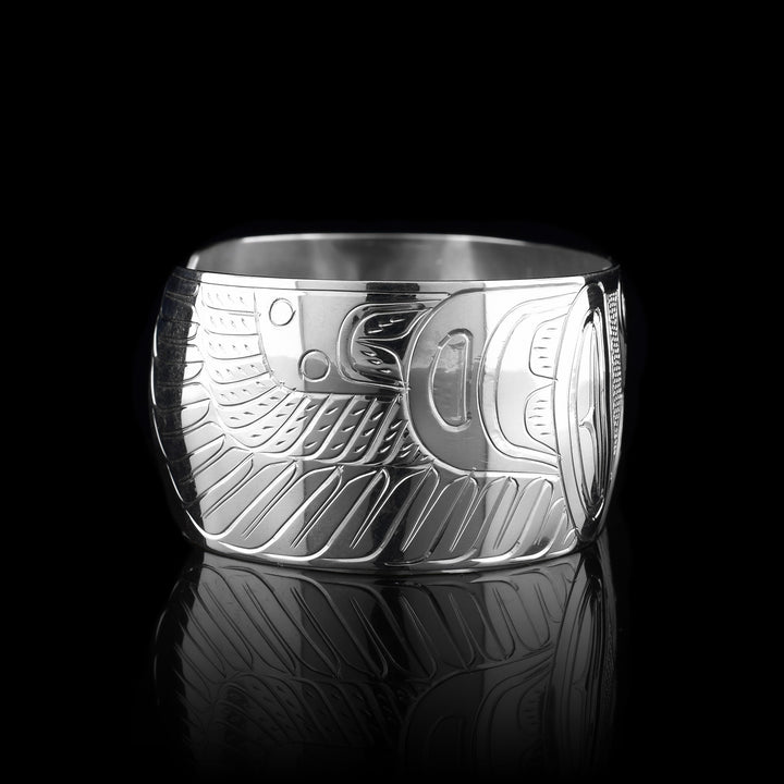 This Thunderbird cuff bracelet has a single, wide band made from sterling silver with a gap in the back. The head of the Thunderbird is shown in the center of the band at a side profile and on either side, its wings are outstretched across the band with feather designs detailed.