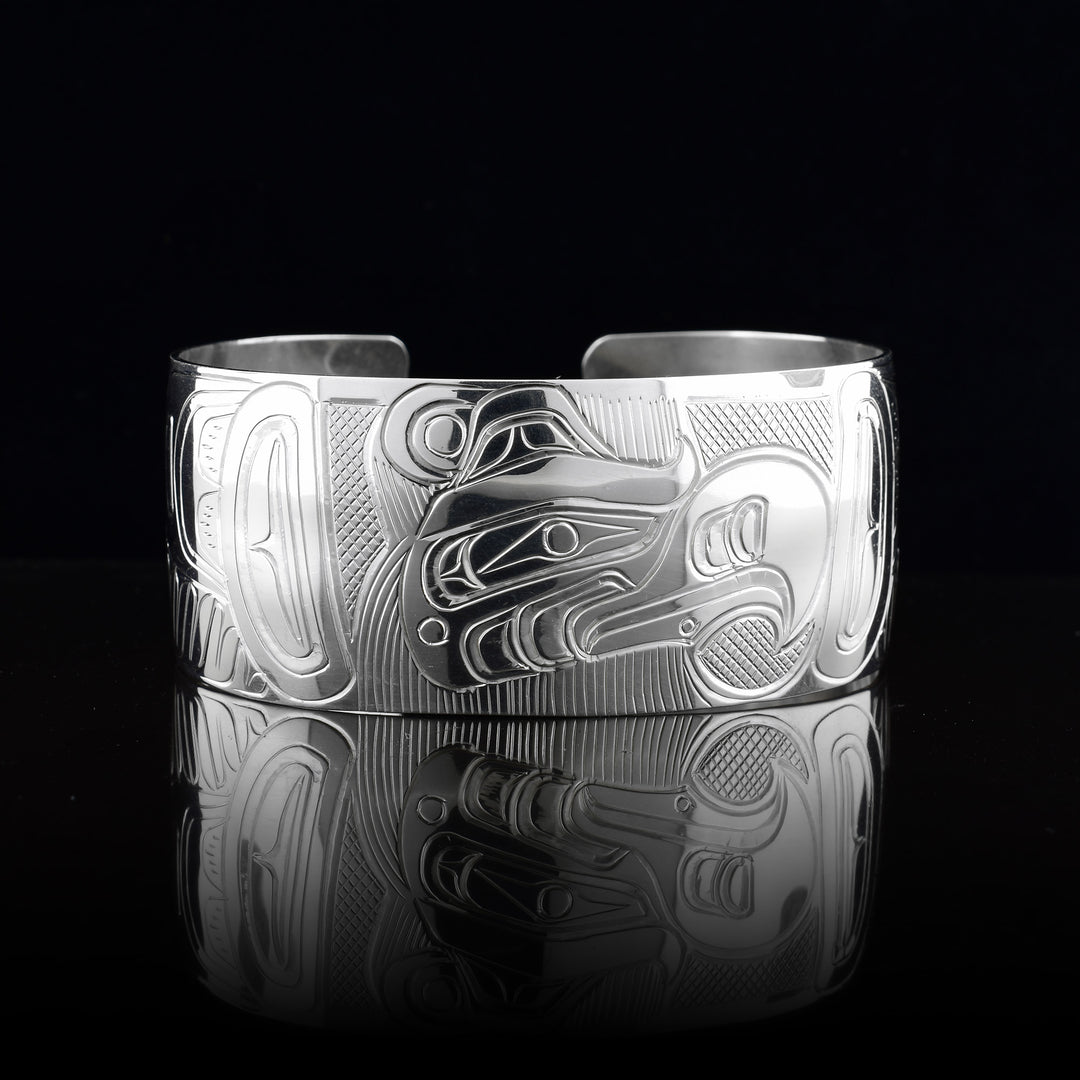 This Thunderbird cuff bracelet has a single, wide band made from sterling silver with a gap in the back. The head of the Thunderbird is shown in the center of the band at a side profile and on either side, its wings are outstretched across the band with feather designs detailed.