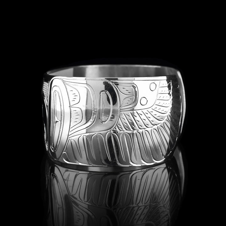 This Thunderbird cuff bracelet has a single, wide band made from sterling silver with a gap in the back. The head of the Thunderbird is shown in the center of the band at a side profile and on either side, its wings are outstretched across the band with feather designs detailed.