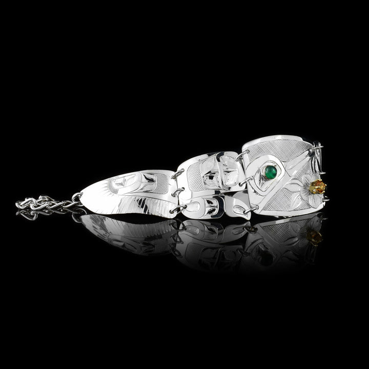 This beautiful panel bracelet has a sterling silver, rectangle panel with a depiction of the Hummingbird and a flower carved into it. There is a green onyx gem in the eye of the legend and a citrine gem in the center of the flower. There are two other panels on either side of the center one that taper. There are designs carved into these panels with the last ones depicting wings of the Hummingbird.