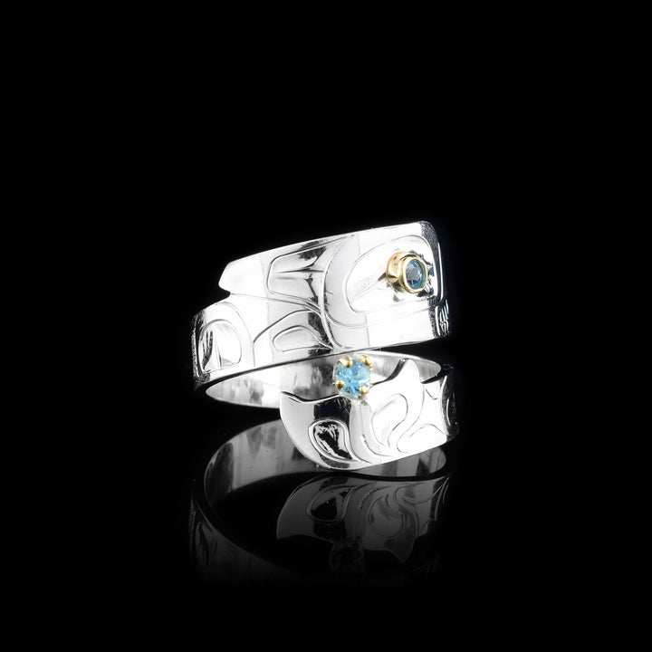This blue topaz ring has a sterling silver band that wraps around the finger twice. The piece depicts the Orca and there are details of the legend carved into the band. The head is at the top with a blue topaz gem set in the eye and the bottom is the tail with another gem held in it.