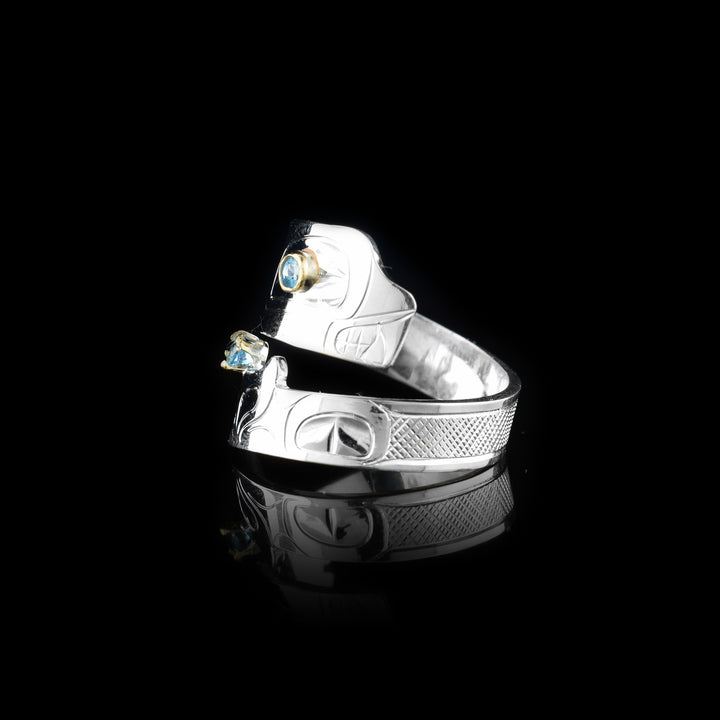 This blue topaz ring has a sterling silver band that wraps around the finger twice. The piece depicts the Orca and there are details of the legend carved into the band. The head is at the top with a blue topaz gem set in the eye and the bottom is the tail with another gem held in it.