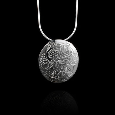 This sterling silver Bear pendant has a round, slightly oblong, shape and there is a depiction of the Bear legend carved into it. The Bear's full body is depicted in a side view and it is sitting with its head up. There are fur details, ears, teeth, eyes and claws included in the scene.