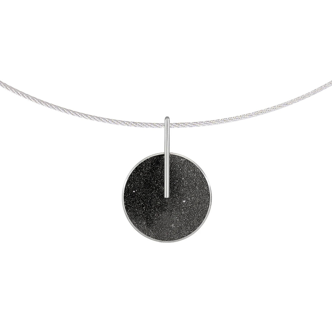 This stainless steel necklace features a large, round pendant made from black-tinted concrete and diamond dust attached to a long, skinny post that connects the pendant to the steel chain.