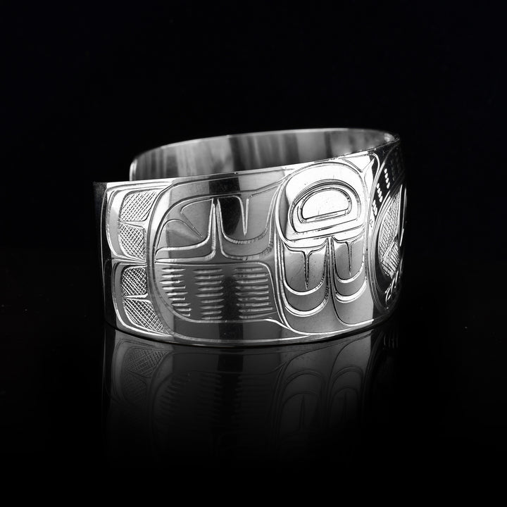 This bracelet for baby has a single, wide band with a gap in the back that is made from sterling silver. The Thunderbird legend is carved at a side profile across the band. The head is large and in the centre of the band. On either side of the head, there are designs representing wings. There are cross hatching designs made in the negative space of the scene.