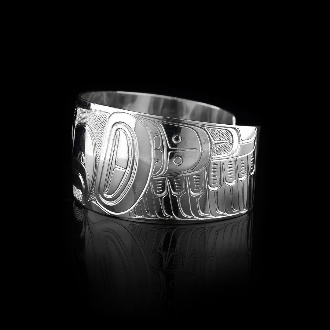 This bracelet for baby has a single, wide band with a gap in the back that is made from sterling silver. The Thunderbird legend is carved at a side profile across the band. The head is large and in the centre of the band. On either side of the head, there are designs representing wings. There are cross hatching designs made in the negative space of the scene.