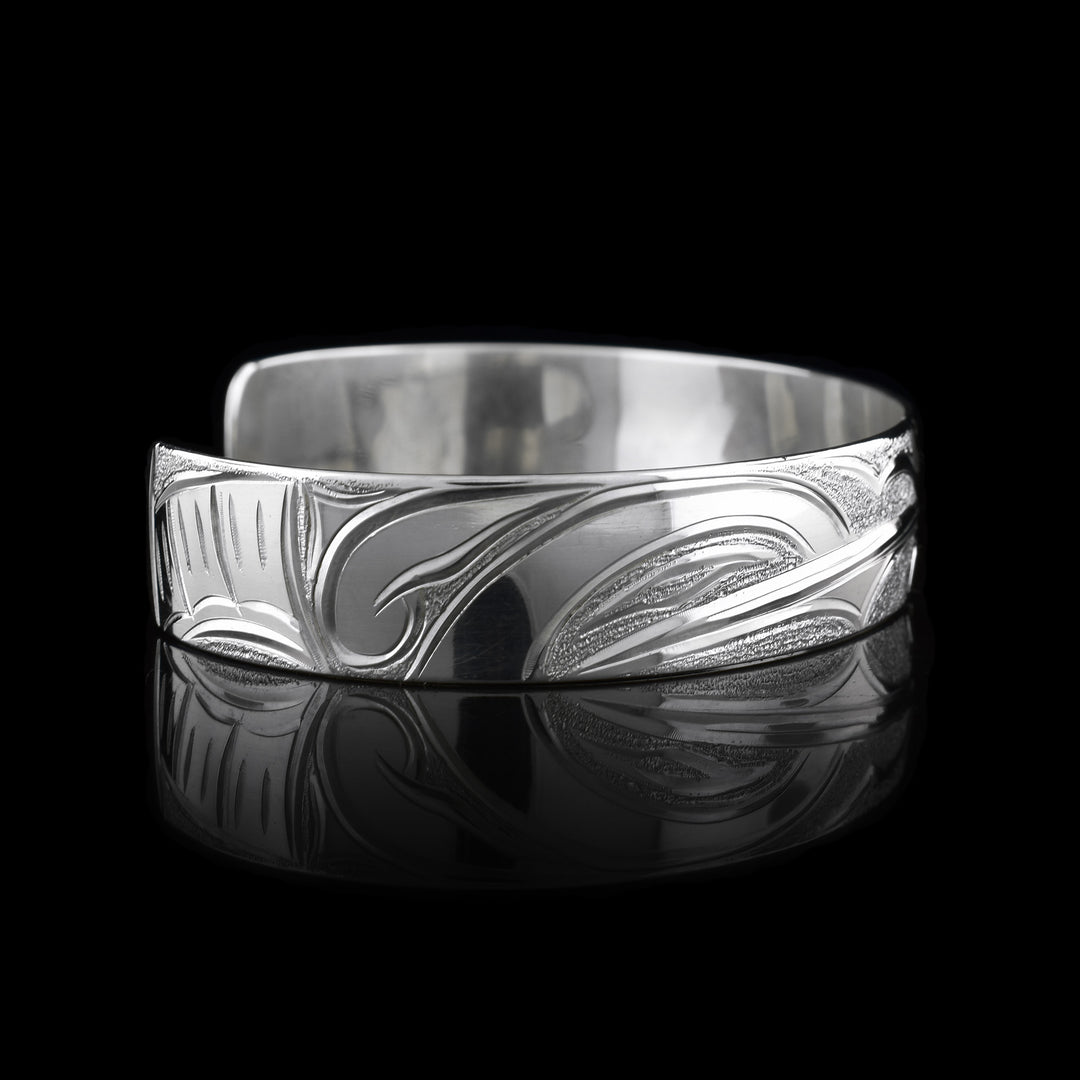 This Hummingbird flower bracelet has a single band with a gap in the back and is made from sterling silver. There are depictions of the Hummingbird legends done in a side profile. There is a flower in the center of the piece and a Hummingbird on either side of the flower. There are dimpled designs in spaces of the scene to give contrast.