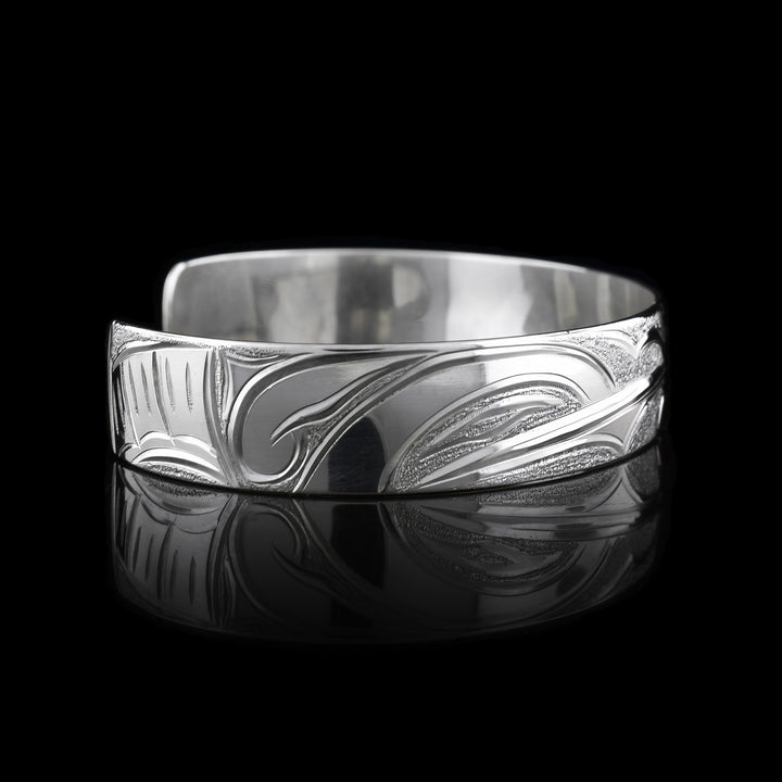 This Hummingbird flower bracelet has a single band with a gap in the back and is made from sterling silver. There are depictions of the Hummingbird legends done in a side profile. There is a flower in the center of the piece and a Hummingbird on either side of the flower. There are dimpled designs in spaces of the scene to give contrast.