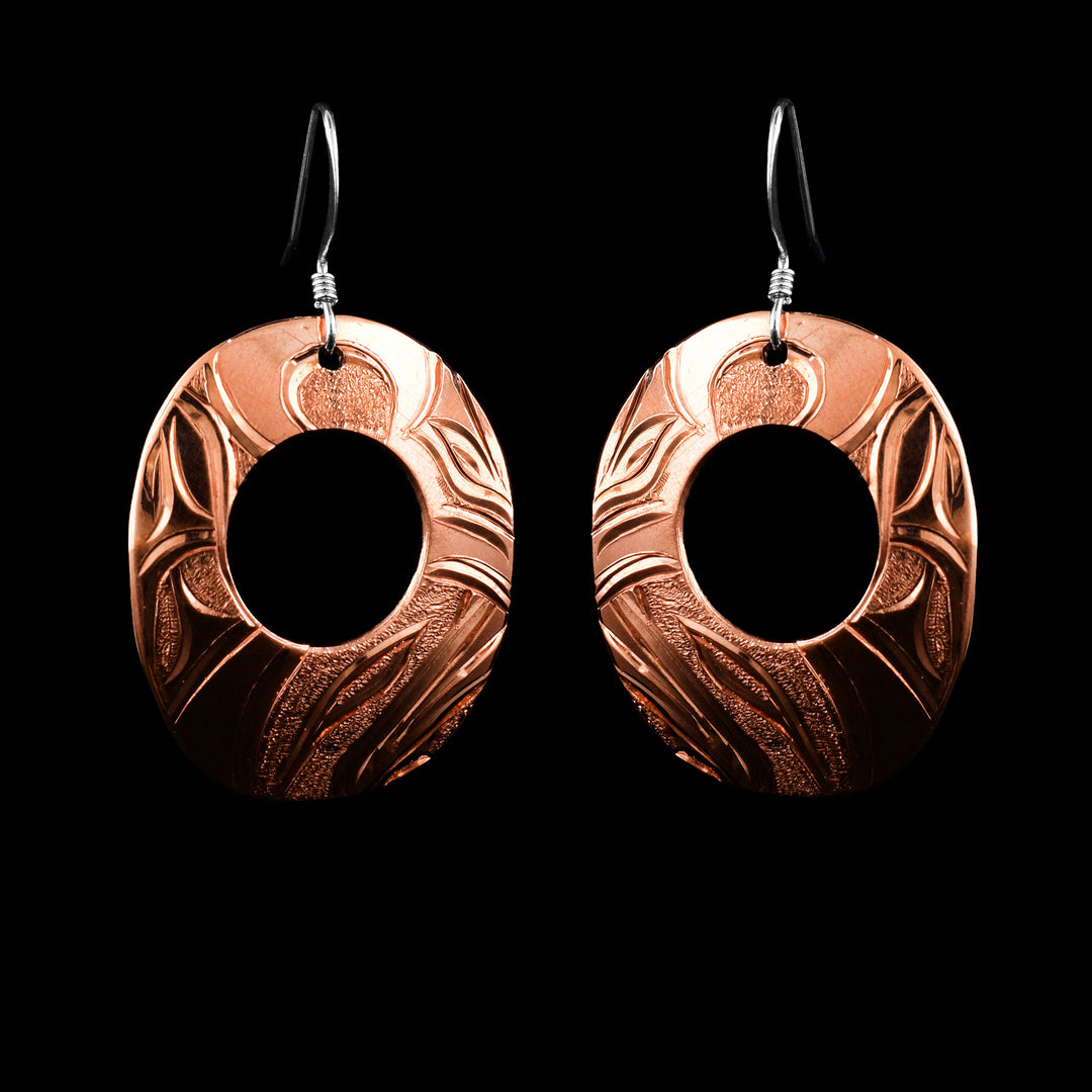 These Orca whale earrings have oval shaped hangs made from copper. The hangs are wide ovals with small cutouts in the center of them. The head of the Orca is carved into the bottom of the hangs and there are wavy designs on the rest of the piece. There are small, dimpled textures in the negative spaces of the scene.