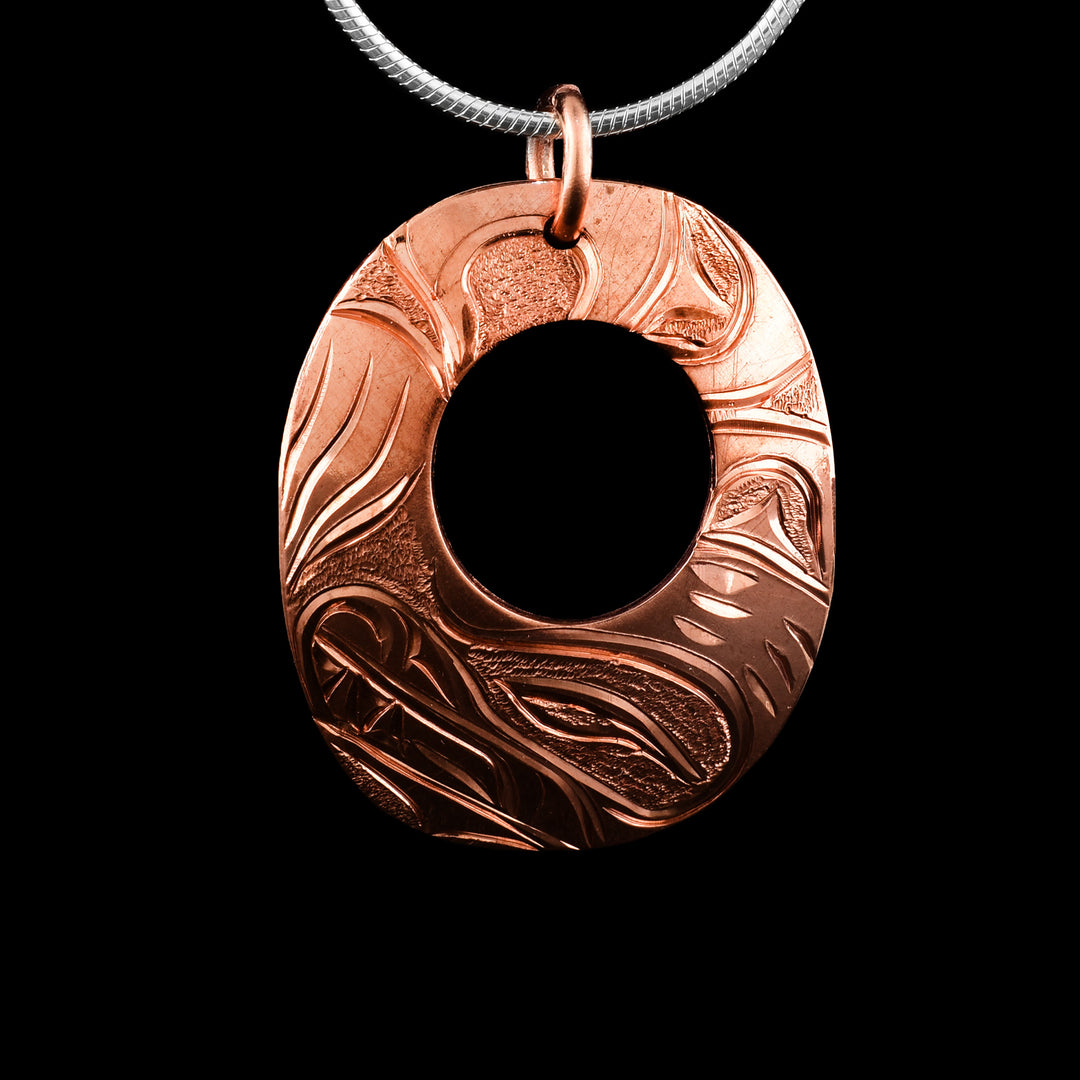 This Bear pendant was a wide, oval shape and a smaller, circle cutout in the center of it. The piece is made from copper and there is a depiction of the Bear legend carved into the surface of it. The head of the Bear is at the bottom of the piece and its clawed, paw is on the left side. Its ears are depicted on the right side of the piece and there are dimpled textures in the negative spaces of the piece.